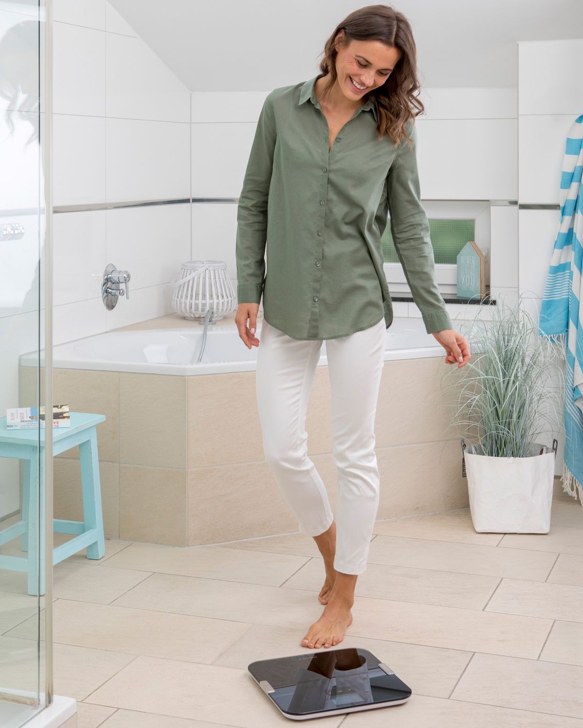 Eine lächelnde Frau steht barfuß im Badezimmer auf einer schwarzen Körperanalysewaage.